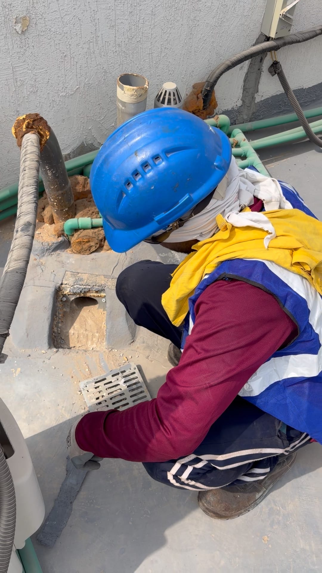 A crew member in a hard hat, hi-vis vest, gloves and safety boots clearing a roof drain, Ajman