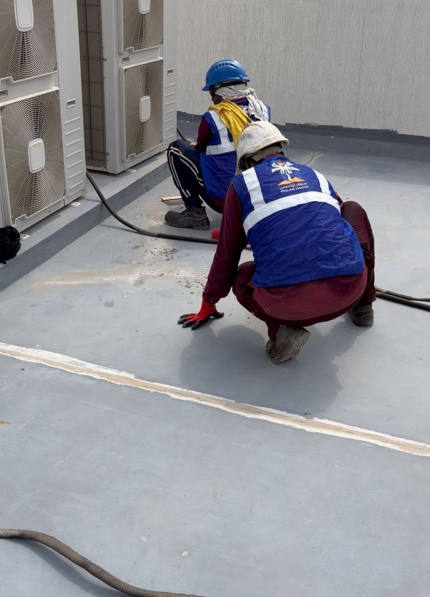 Grey villa roof before the cool-roof coating, with two Solar Oasis crew in hard hats preparing the surface beside a sealed joint, Ajman