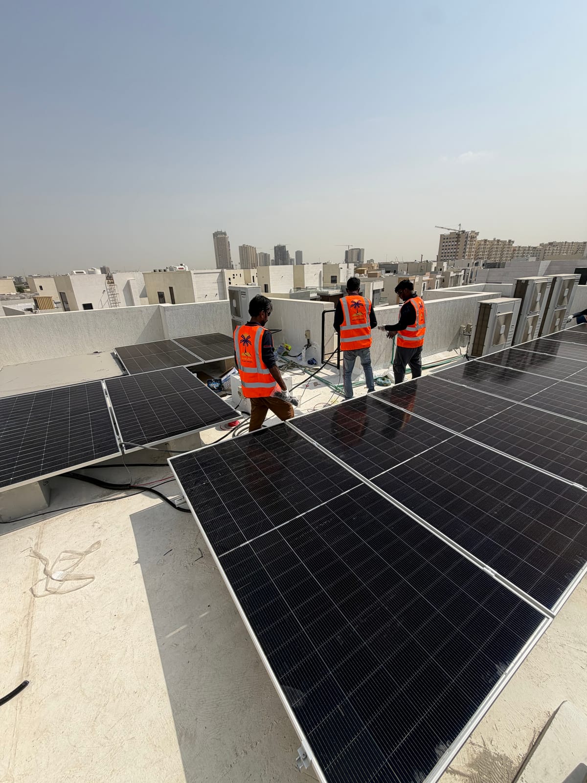 Three Solar Oasis crew in palm-logo hi-vis vests at the parapet of a villa roof, beside rows of solar panels, Ajman