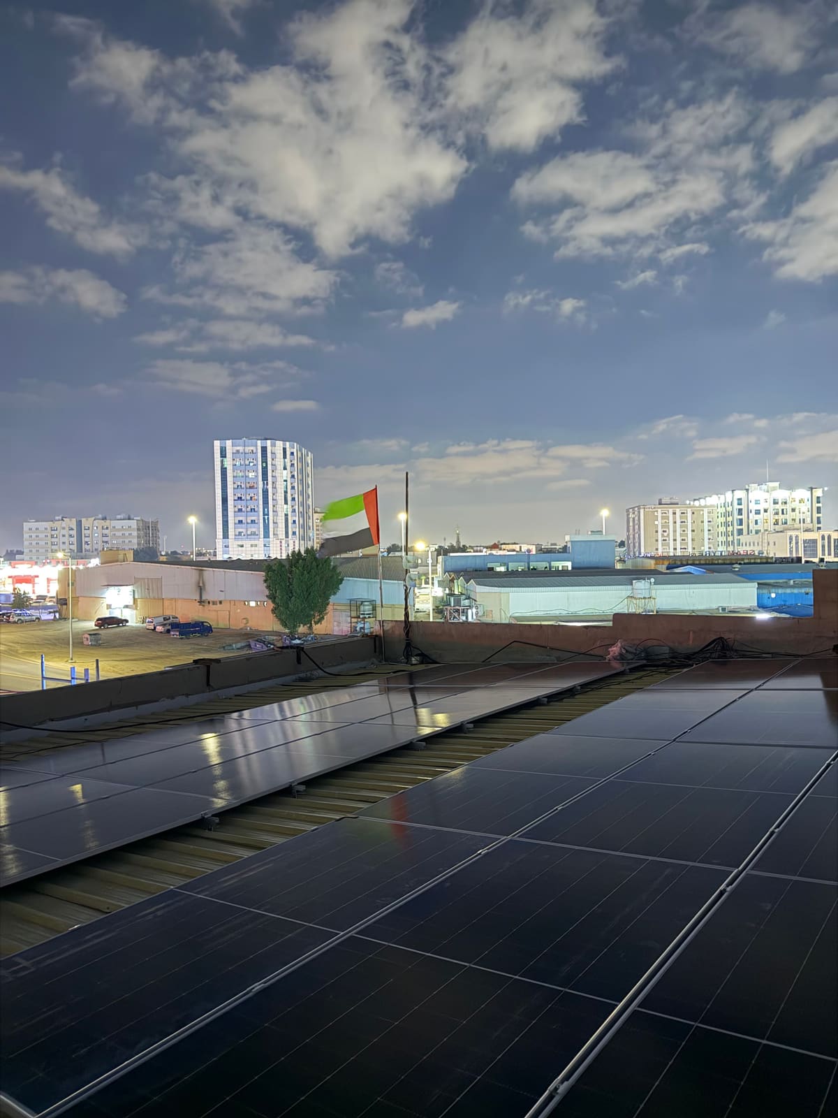 Solar panels on the sandwich-panel roof of Bu Faisal General Trading at dusk, with the UAE flag flying behind, Al Jurf, Ajman